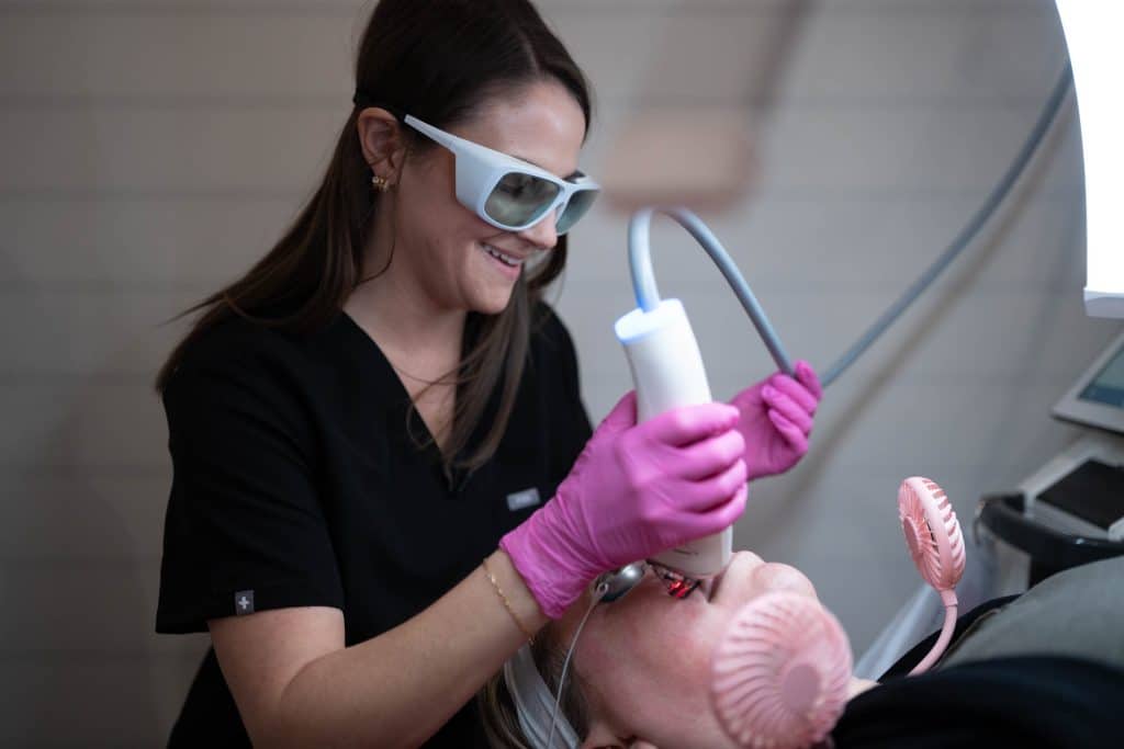 Dental assistant in pink gloves uses a suction device beside a patient’s open mouth during a dental procedure, wearing protective eyewear and black scrubs.