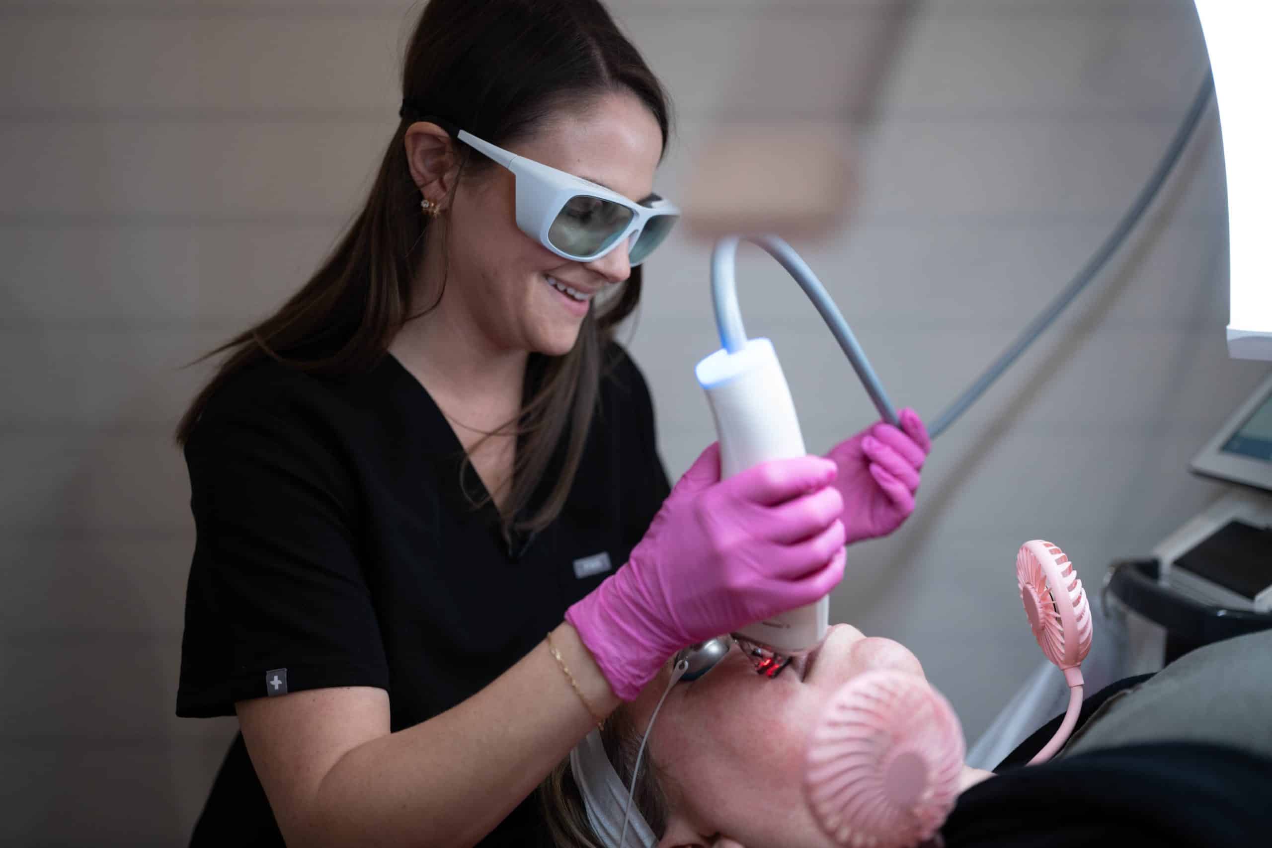 Dental assistant in pink gloves uses a suction device beside a patient’s open mouth during a dental procedure, wearing protective eyewear and black scrubs.