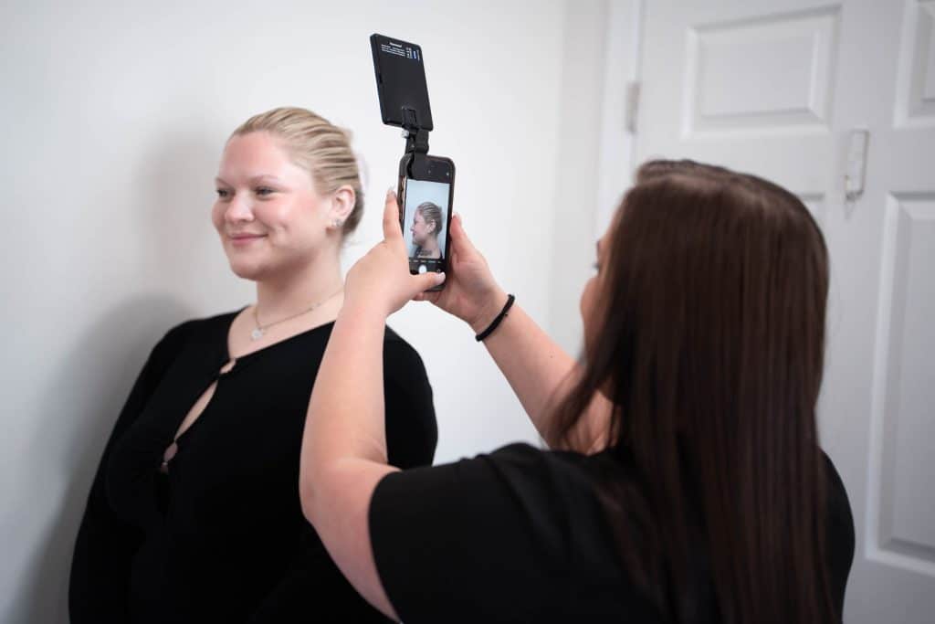 Woman in a black top smiles as she is filmed with a phone mounted on a handheld stabilizer against a light wall behind her.